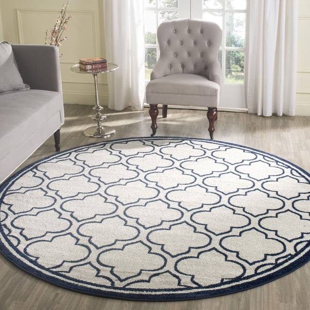 Safavieh Amherst Wilson Geometric Area Rug, a round indoor-outdoor polypropylene rug with a white base and navy blue geometric pattern, displayed on light wood flooring in a bright room with a grey tufted accent chair, grey sofa, and glass side table with books and a floral branch, ideal for high-traffic home or office spaces.