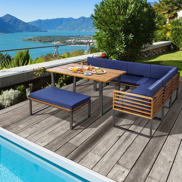 Gymax 8PCS Patio Dining Set with acacia wood frame and navy cushions, featuring an L-shaped sectional sofa, dining table, ottoman, and corner and armless sofas. Ideal for outdoor dining or relaxing on a patio, deck, or poolside. Set shown on a wooden deck overlooking a lake and mountains.