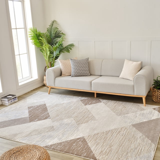 Rug Branch Nomad Abstract Indoor Area Rug in beige and brown, 4'x6' (3'11" x 5'7"), featuring geometric abstract patterns in textured polypropylene. Displayed in a modern living room with a light gray sofa, wooden legs, and three patterned pillows, alongside a tall green plant and woven baskets on a light wood floor, suitable for living, dining, or kitchen spaces.