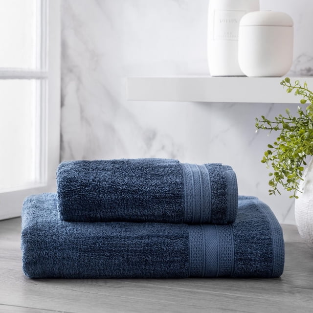 hometrends Solid Bath Towel in deep blue, featuring two folded towels with textured bands stacked on a light wood surface near a window, set against a marble wall with white bathroom accessories and a green plant, made from 100% cotton for softness and high absorbency, ideal for bathroom use and spa-like comfort.