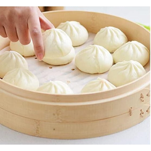 A close-up of a bamboo steamer basket with white steamed buns resting on perforated parchment paper, with a hand picking up one bun, demonstrating the use of a food liner ideal for steaming and baking.