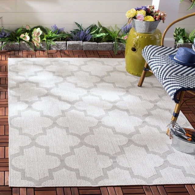 SAFAVIEH Bermuda Sophie Geometric Machine Washable Indoor/Outdoor Area Rug in a light neutral tone with a repeating geometric quatrefoil pattern and textured weave, placed on a wooden deck patio. This weather-resistant, stain-resistant rug is ideal for outdoor living spaces, shown with garden plants, a patterned chair, and a green ceramic side table holding a metal bucket of flowers.