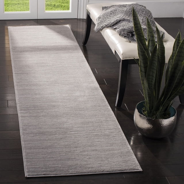 Safavieh Vision Adrasteia Overdyed Solid Area Rug in a soft light neutral tone with subtle linear texture, laid on dark hardwood flooring beside a white leather bench draped with a gray knitted throw and a tall snake plant in a hammered silver pot near glass doors revealing green grass outside, suitable for modern hallways or living rooms.