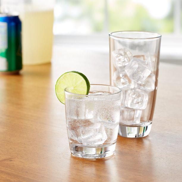 Mainstays Glassware 16-piece set with clear, tapered glass design and sturdy base, featuring a 9.7-oz rock glass with ice and lime slice, and a 15.8-oz cooler glass with ice on a wooden table, ideal for water, soda, or mixed drinks.