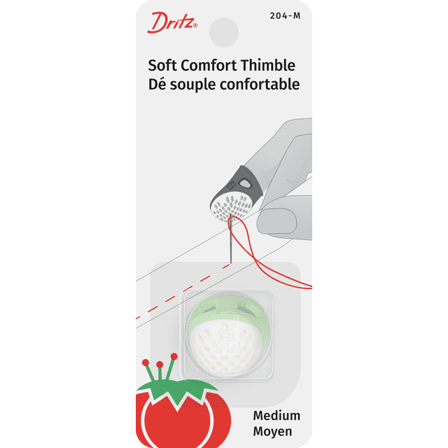 Dritz Soft Comfort Thimble medium green with white dimpled tip worn on finger pushing a needle threaded with red thread through fabric, featuring ergonomic design for needle protection, shown on packaging card with bilingual text and tomato pin cushion graphic, also suitable for quilting and crafting projects.