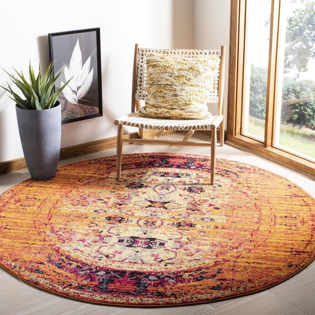 Safavieh Monaco Elsdon Abstract Area Rug in a bright corner featuring a round rug with a Bohemian-chic abstract Persian pattern in warm orange, red, and cream colours. The durable polypropylene rug is power-loomed with mixed high and low loop pile. It is styled with a woven wooden chair holding a yellow and white textured cushion, a gray planter with a green succulent, and a framed leaf print on the wall. This rug is also suitable for adding vibrant style to both country and city homes.