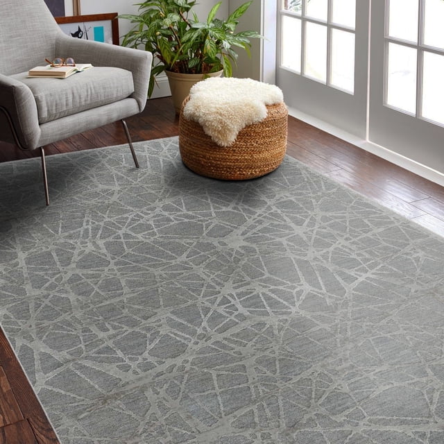 Rug Branch Melo Collection Modern Abstract Scandinavian Indoor Area Rug with a rectangular grey surface and lighter grey abstract intersecting lines, placed on dark hardwood floors in a bright room featuring a grey armchair, woven round ottoman with white sheepskin, and a green potted plant near white French doors. Made of 100% polyester with jute backing, suitable for living rooms and hallways.