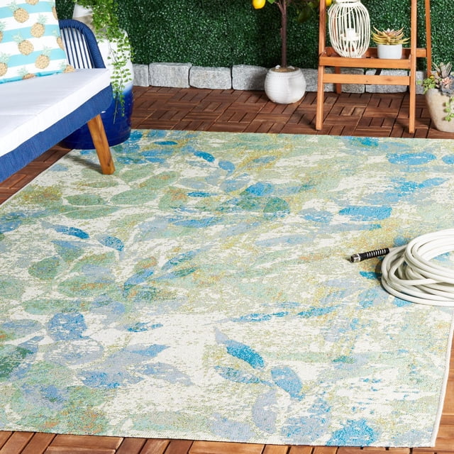 SAFAVIEH Barbados Gresham Tapis Botanique Intérieur/extérieur avec Motif de Feuilles Botaniques dans des Tons Bleu, Vert et beige, Posé sur un Plancher de patio en Bois à Côté d'Une Chaise Rembourrée Bleue avec des Coussins à Motif d'Ananas, un Tuyau d'Arrosage Blanc Enroulé et des Plantes en Pot sur un Fond de Haie Verte, Convenant à une Déco Extérieure Vibrante.