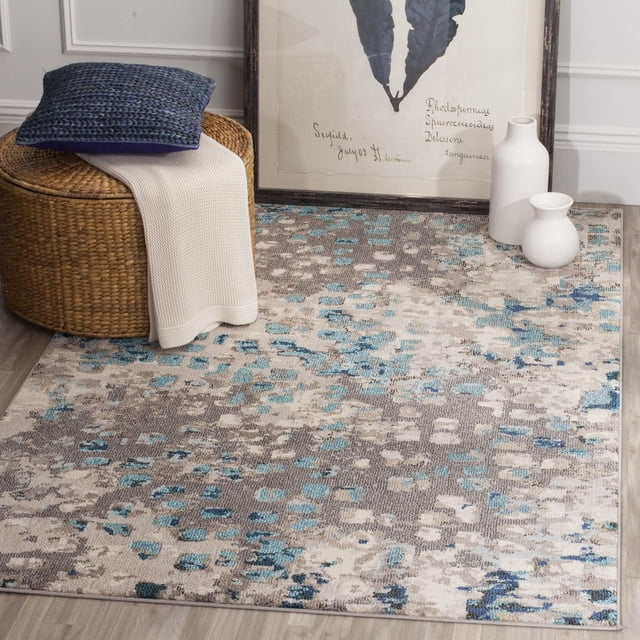 Safavieh Monaco Driskoll Tapis Abstrait avec un Motif Abstrait Pixélisé en Polypropylène Bleu, Gris et beige Empilé sur un Plancher de Bois Clair. le Tapis Est Conçu avec un ottoman Rond Tissé Tenant un Coussin Bleu et un Jeté Crème, à Côté de vases en Céramique Blancs et d'Un Imprimé Botanique Encadré contre un Mur Blanc. Idéal pour Ajouter une Touche Bohème-Chic aux Espaces de Tapis d'Accent.
