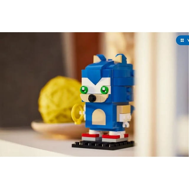 LEGO BrickHeadz Sonic The Hedgehog (40627) set featuring a blocky, stylized version of Sonic the Hedgehog with blue and beige bricks, green eyes, and red shoes. The figure is displayed on a black baseplate and includes a yellow ring accessory. It stands over 3 inches (8 cm) tall and is shown on a shelf with a blurred yellow decorative object in the background.