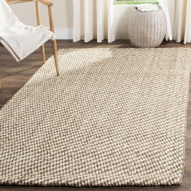 Safavieh Natural Fiber Daisy Braided Area Rug in natural beige with a textured braided sisal weave, placed on dark hardwood flooring beside a wooden chair with a white throw and a round woven side table near a window with white curtains, suitable for coastal and casual interiors.
