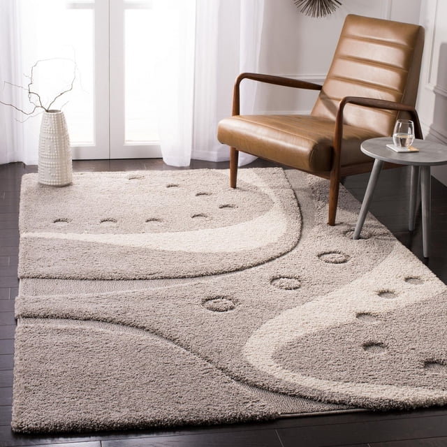 Safavieh Florida Laidley Contemporary Shag Area Rug featuring a plush crème tone-on-tone high-low pile with damask vine patterns, placed on dark hardwood flooring beside a tan leather armchair and a small round side table with a glass of water, suitable for enhancing texture and comfort in living rooms.