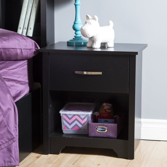 South Shore Fusion 1-Drawer Nightstand in pure black with a metal handle, featuring a deep drawer and open storage space holding a plastic container and purple fabric bin, topped with a blue lamp and white ceramic dog figurine, placed beside a bed with purple bedding on a light wood floor and gray wall.