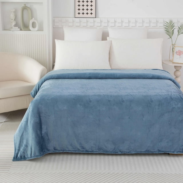 Mainstays Kids Embossed Blanket Gamer in blue polyester blend spread over a bed with white pillows in a bright, minimalist bedroom featuring a cream armchair, geometric wall art, and a round side table with a plant and flamingo print, ideal for warmth and playful use like blanket forts.