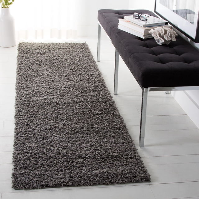 Safavieh Athens Solid Plush Shag Area Rug in grey, displayed as a rectangular runner on white wood flooring beside a black tufted bench with chrome legs, featuring a soft, dense polypropylene pile that complements both modern and traditional interiors and is ideal for adding warmth and texture to hallways or entryways.