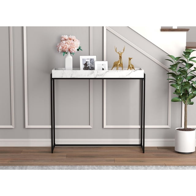Safdie & Co. Console Table with white marble-look MDF removable tray top and black metal legs, styled with a white vase holding pink flowers, two gold deer figurines, and two black-and-white photo frames, set against a light grey wall with decorative molding, wood flooring, and a potted plant near a staircase.