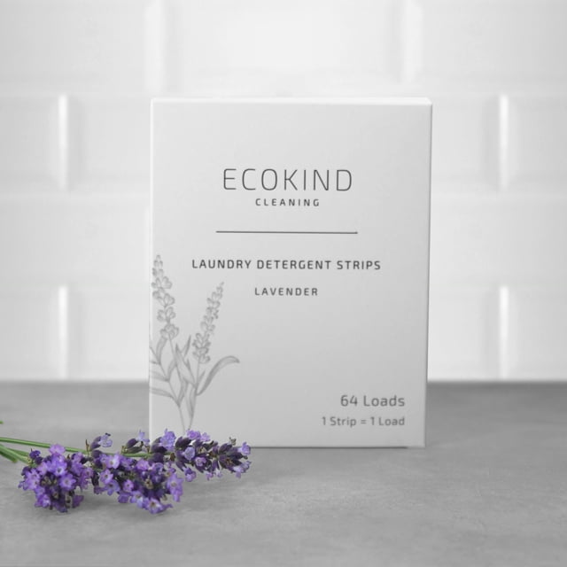 ECOKIND Cleaning Laundry Detergent Strips in lavender scent, displayed in a white box with lavender illustrations. The packaging notes 64 loads per pack and emphasizes eco-friendly, plant-based ingredients. A sprig of lavender lies on a grey surface in the foreground, with a white tiled background.