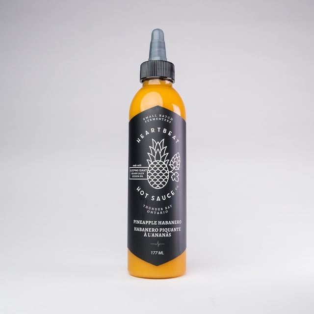 Sauce Piquante Habanero à l'Ananas en Bouteille Transparente Orientée vers l'Avant avec Ambre-Orange, Sauce Versable Mettant en Valeur des Taches Rouges de Habanero et des Particules d'Ananas; l'Étiquette Lecture une Sauce Piquante au Battement de Cœur, Fabriquée au Canada, Petit Lot, Végane, Sans Agent de Conservation. à l'Avant-fruit avec des notes de lime Vives, Chauffe 5/10, Idéale sur tacos, pizza et Poulet et Souvent Utilisée comme base de marinade.