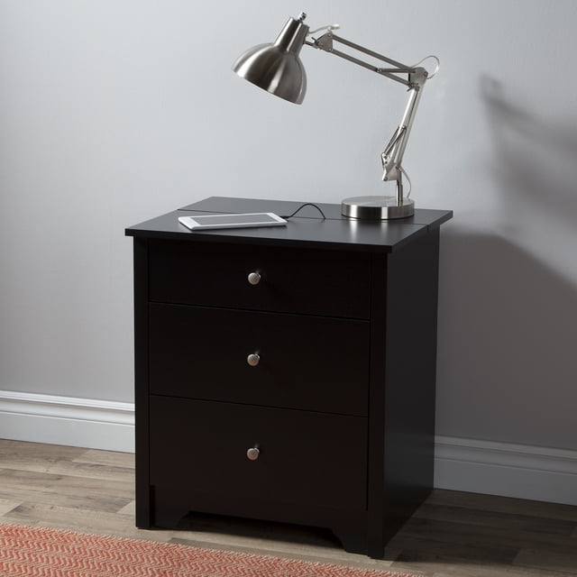 South Shore Vito Nightstand with Charging Station and Drawers for Adult in pure black, front view showing three drawers with metal knobs, a sturdy top with a sliding tray charging station holding a silver adjustable desk lamp and a tablet plugged in, set against a light grey wall on wood flooring with a red patterned rug edge visible, ideal for organizing bedroom essentials and charging devices neatly.