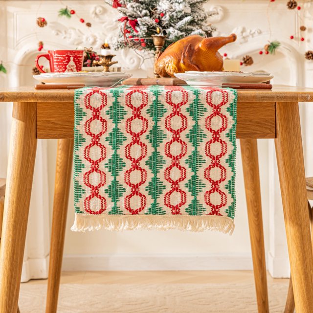 Christmas Boho Macrame Table Runner, 72 inches long, displayed on a wooden dining table. The runner features a woven design with red and green patterns resembling Christmas trees and geometric shapes, complemented by fringe detailing on both ends. The table is set with festive decor, including a roasted turkey, red holiday-themed mug, and candles, creating a cozy and elegant Christmas atmosphere. Made of 85% cotton and 15% linen, this rustic table runner is suitable for indoor and outdoor holiday gatherings, enhancing farmhouse and vintage decor styles.