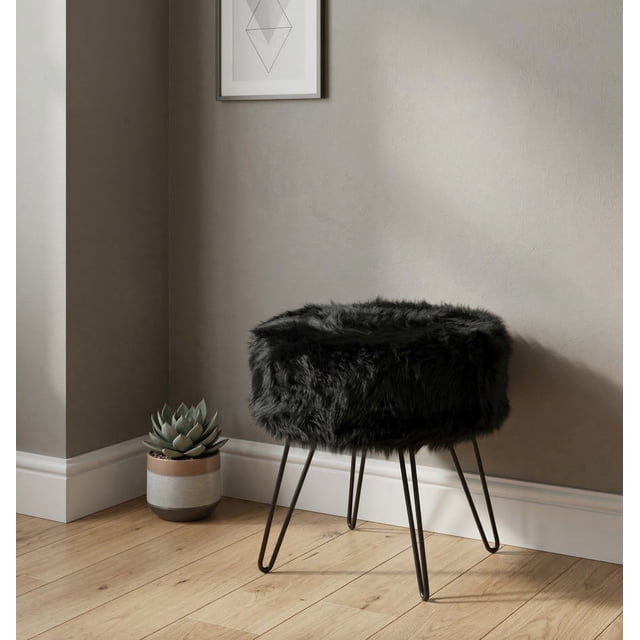 Ottoman Rond Pixie, Pouf, Repose-Pieds, Tabouret Repose-Pieds avec un design Luxueux en Fausse Fourrure Noire et des Pattes en Épingle à Cheveux en Métal Noir. l'Ottoman Présente un Siège Rond avec un Diamètre de 14,5 Pouces et une Hauteur d'Assise de 16 Pouces. construction Robuste Supporte jusqu'à 300 Lb. Exposé dans un Décor de Salon Moderne avec un Mur Gris Pâle, un Plancher en Bois et une Petite Plante en Pot à Proximité.