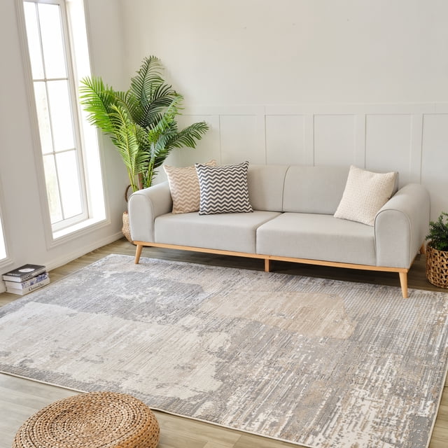 Rug Branch Nomad Abstract Indoor Area Rug in cream grey with a modern abstract pattern, displayed in a bright living room with a light grey sofa with wooden legs and three patterned cushions, a tall green potted plant by a large window, and a woven round ottoman on light wood flooring, ideal for living room, bedroom, dining room, or kitchen settings.