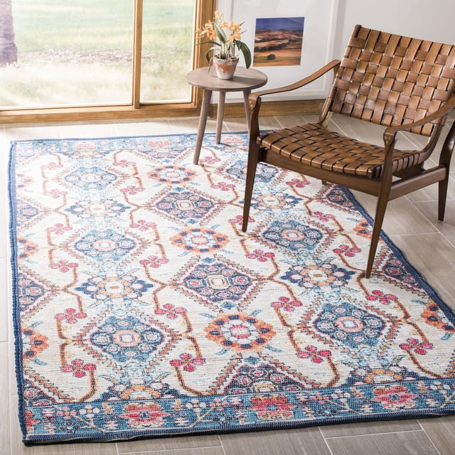 Safavieh Saffron Ira Floral Bordered Area Rug with a hand-made cotton weave displaying a distressed floral and geometric pattern in blue, red, and orange on a cream background, placed on a light wood floor next to a woven leather chair and a round wooden side table with a potted orchid, in a naturally lit room. The rug's soft cotton texture makes it suitable for cozy living spaces or as an accent piece in country or city homes.