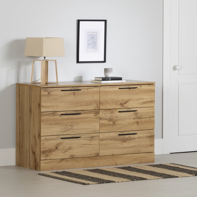 South Shore Step One Essential 6-Drawer Dresser for Adult in Nordik Oak with six drawers featuring black metal handles and metal slides for durable storage. The modern minimalist dresser has a large top surface holding a wooden lamp with beige shade, two books, and a candle, set against a light grey wall with framed abstract art and a striped rug on the floor. It is also suitable for organizing craft or office supplies.