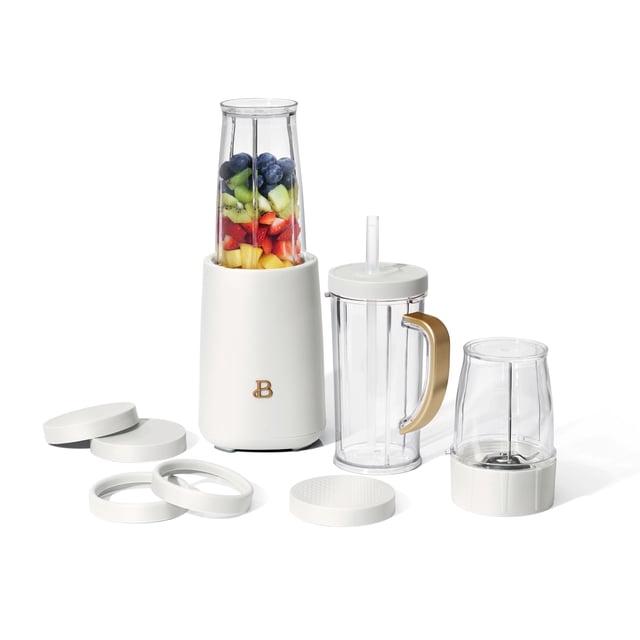Beautiful 12pc Personal Blender by Drew Barrymore with white matte base and gold 'B' logo, tall blending jar filled with fresh fruit, 16oz ToGo travel cup with gold handle and no-drip flexible straw lid, grinding cup with stainless steel blade, and multiple dishwasher-safe lids and lip rings, suitable for smoothies, sauces, and grinding coffee beans.