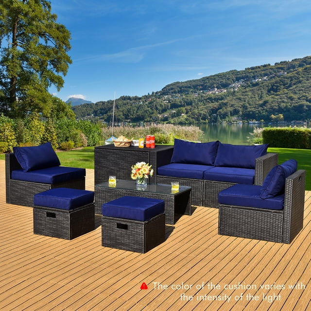 Gymax 8PCS Rattan Patio Sectional Furniture Set with waterproof cover and navy cushions, featuring a black PE rattan frame and ergonomic design. Includes a tempered glass coffee table, storage box, two ottomans, two armless sofas, and two corner sofas. The set is displayed on a wooden deck with a scenic lake and mountain view in the background. Cushions are navy blue, and the set is arranged for outdoor relaxation.