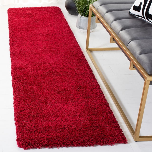 Safavieh Milan Harlow Tapis Shag Uni en Rouge Vif, un Coureur Shag en Peluche Synthétique Exposé sur un Plancher Blanc à Côté d'Un Banc Moderne à Cadre Métallique Doré avec des Coussins Noirs et un Coussin Décoratif, Idéal pour Ajouter Chaleur et style aux Intérieurs Contemporains de la Maison.