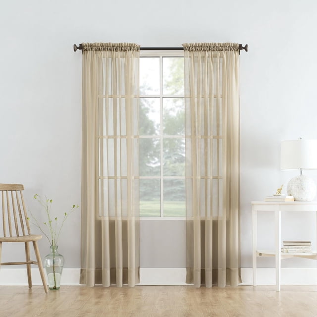 SUN TEXTILE VOILE POLE TOP CURTAIN in beige sheer voile fabric with built-in rod pockets hanging on a dark metal rod over a window with grid panes, gently filtering light and enhancing privacy in a minimalist room with light wood flooring, a wooden chair with a glass vase holding green stems on the left, and a white side table with books and a white lamp on the right, suitable for layering with other window coverings.