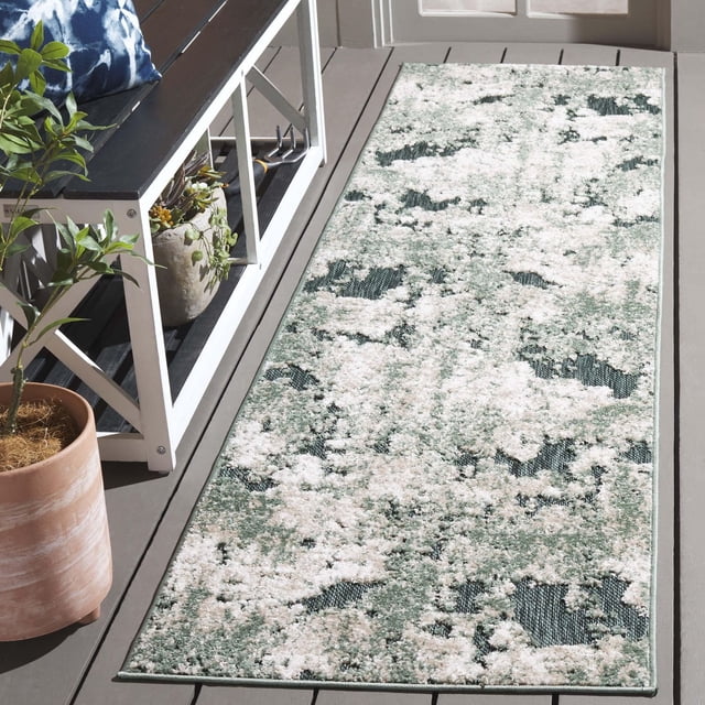 SAFAVIEH Cabana Selma Distressed Indoor/Outdoor Area Rug on a grey wooden deck, featuring a textured floral and geometric pattern in muted green and cream, made with durable synthetic yarns resistant to weather and sun fading, ideal for busy indoor or outdoor areas including porches and patios.