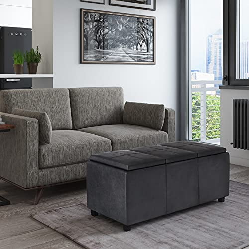 Simpli Home Avalon Rectangle Storage Ottoman with 3 flip over serving trays, upholstered in distressed black faux leather. The ottoman, suitable as a coffee table, is placed in a modern living room setting with a grey sofa and large windows. Features include a stitched exterior, solid wood construction, and spacious interior storage.