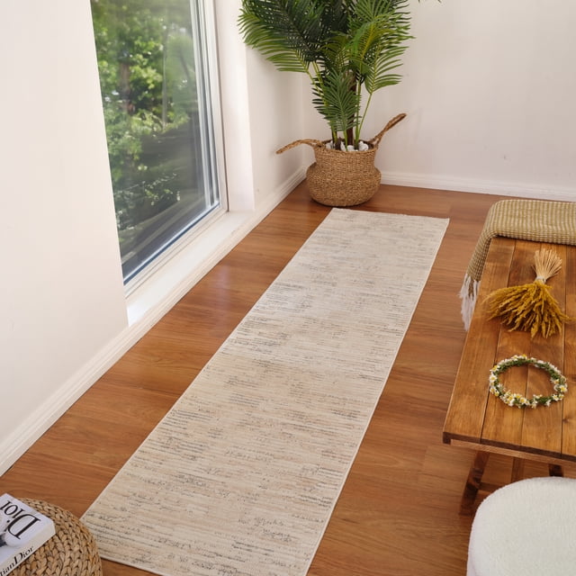 Rug Branch Astra Modern Indoor Area Rug in beige cream with an abstract pattern, shown as a runner on a wooden floor near a large window. Made of easy-care polyester with jute backing, it suits high-traffic areas like kitchens and hallways. The scene includes a woven basket with a green plant and a wooden bench with decor, illustrating its versatile indoor use.