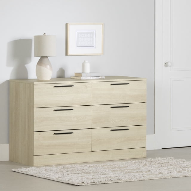 South Shore Step One Essential 6-Drawer Dresser for Adult in bleached oak with six drawers featuring black metal handles and metal slides, set against a light grey wall with a framed abstract print above, topped with a beige ceramic lamp, books, and a white vase, placed on a light textured rug near a white door, ideal for organized bedroom storage.