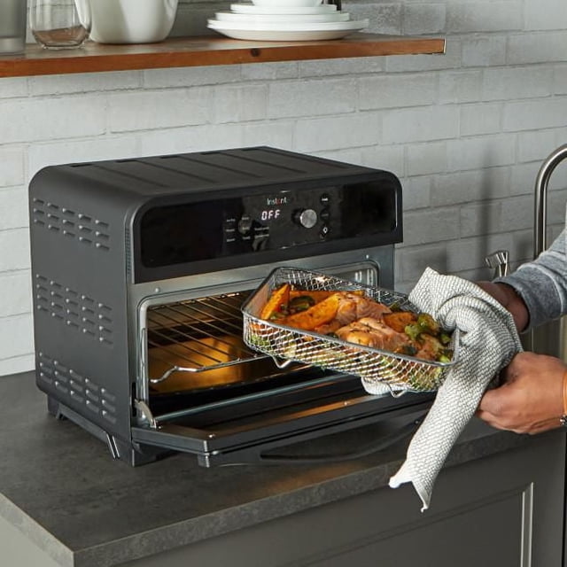 Instant® Omni 18L 7-in-1 Air Fryer Toaster Oven. Centered three-quarter countertop view of the stainless-steel oven with front glass door and digital display; enamel baking tray, air-fry basket and stainless-steel rack visible inside the cavity; control knob, LED panel, black handle and side vent slots visible; capacity fits a 12-inch pizza (18 L) and control panel highlights convection and EvenCrisp technology; often used for reheating or crisping meal-prep portions.