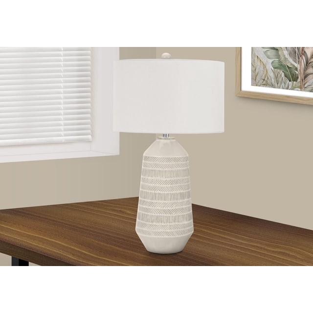 Monarch Specialties Lighting 33-inch contemporary table lamp with a cream ceramic textured base featuring horizontal carved patterns, topped with an ivory fabric drum shade and round ceramic finial, placed on a wooden side table near a window with white blinds and framed botanical artwork on a beige wall, ideal for bedroom or living room use.