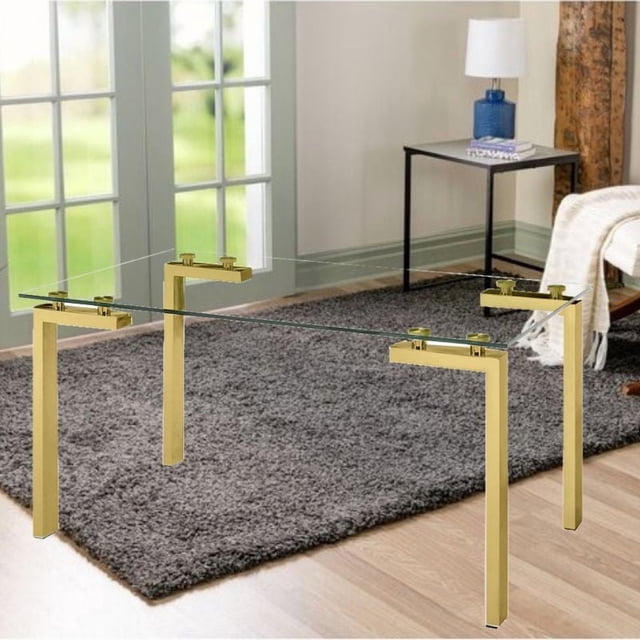 Emma Dining Table in Gold with a clear tempered glass rectangular top supported by four gold stainless steel legs, set on a gray rug over wooden flooring in a bright room with a side table and blue lamp, suitable for dining or kitchen use.