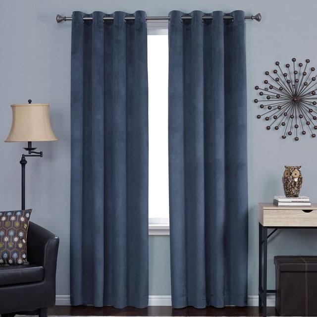Hometrends Velvet 84" Grommet Blackout Window Curtains Panel in indigo blue velvet with metal grommet top, hanging on a silver rod in a living room. The blackout panel reduces light, noise, and drafts while providing privacy and energy savings. Floor-length and pleated, shown with a black leather chair, beige lamp, and side table with decor.