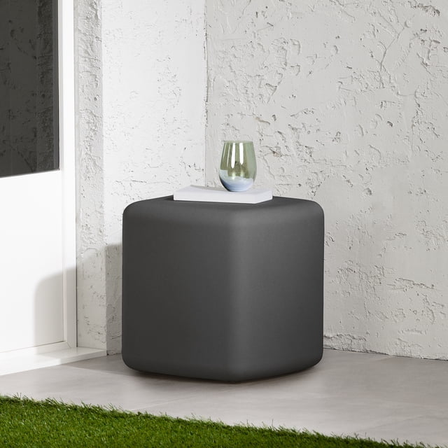 South Shore Dalya Outdoor Side Table with Rounded Corners in dark gray, front-facing on a tiled patio floor against a textured white wall. The compact cubic table has smooth rounded edges and a matte polyethylene resin surface. A white book and green glass rest on top. It resists UV rays, water, and scratches, suitable for small balconies or patios, and can also serve as extra seating.