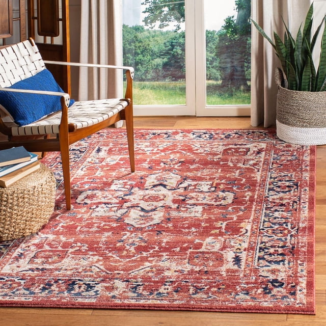 SAFAVIEH Charleston Zharko Overdyed Floral Area Rug with traditional floral patterns in warm red and navy tones, placed on hardwood flooring in a bright room featuring a woven white strap wooden accent chair with blue cushion, a woven ottoman with books, and a large snake plant in a woven basket near glass doors overlooking greenery, ideal for living or dining rooms.