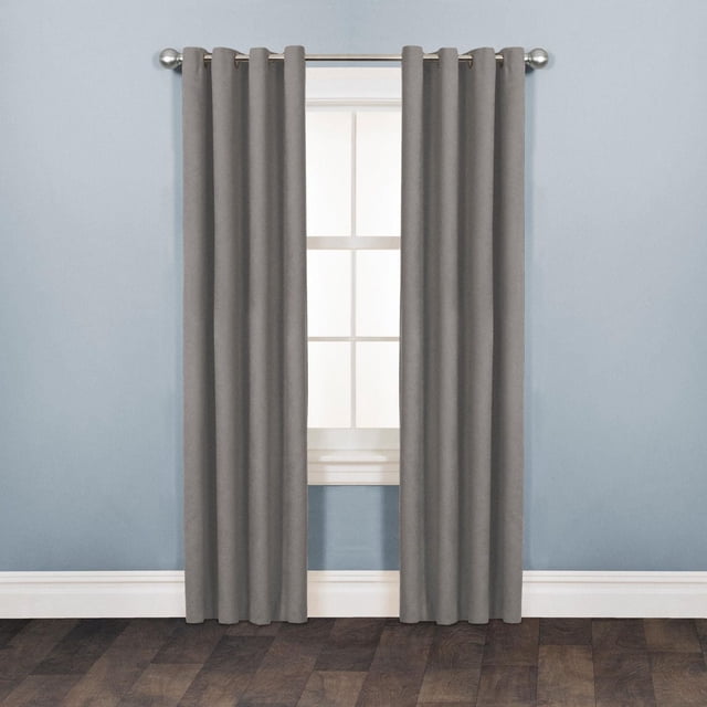 Mainstays Faux Suede Drapery Grommet Window Curtains Panel in matte grey, hanging on a silver curtain rod with metal grommets, covering a white-framed window in a room with light blue walls and dark wood flooring. The 100% polyester panel features a soft suede texture and is designed for easy installation and room darkening, also suitable for adding warmth and style to living spaces.
