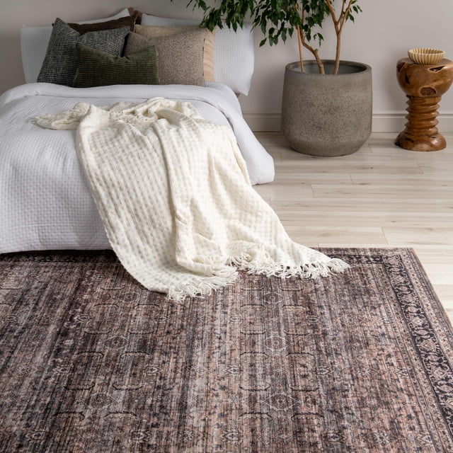 Rug Branch Emir Collection Tapis Traditionnel Oriental Usé Hydrofuge Présentant un Motif Oriental Usé Brun et Gris Atténué avec des Bordures Détaillées, Posé sur un Plancher de Bois Clair dans une Chambre. le Tapis Complète un Couvre-Lit Texturé Blanc, des Coussins Neutres et un Jeté à Franges Crème. à Proximité Se Trouvent un Grand Pot Gris avec une Plante Verte et un Tabouret Décoratif en Bois. Ce Tapis en polyester durable Est Idéal pour les Zones à Fort Passage et Ajoute un style Classique aux Espaces de Vie.