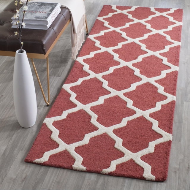 Safavieh Cambridge Liam Geometric Area Rug in red with white geometric lattice pattern, hand-tufted wool with plush cut and high/low loop pile, displayed on light grey wood floor beside a brown leather bench and white vase with flowers, ideal for adding modern American style to home or office.