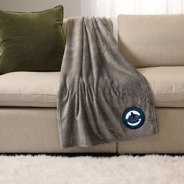 NHL, NBA, MLB Grey Sports Throw Blanket, 50" x 60", draped over a beige sofa with a green cushion, made of soft plush 100% polyester fabric, featuring a Vancouver Canucks logo patch, ideal for indoor warmth and lounging.