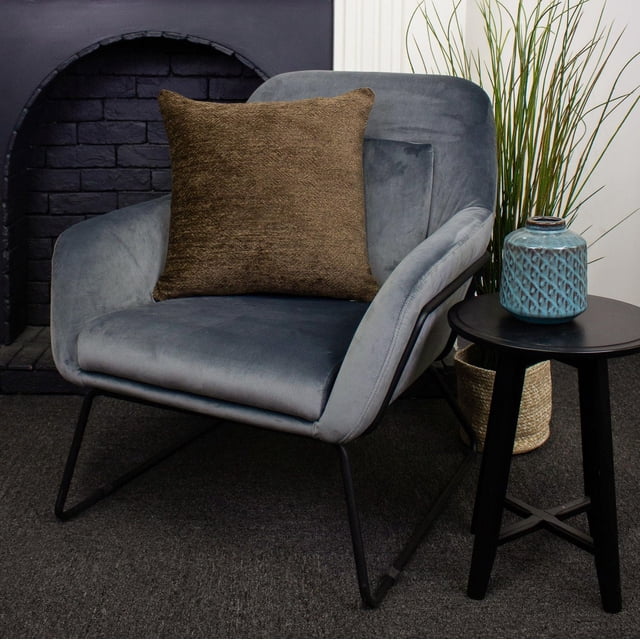Julep Coussin de Luxe en Tissu polyester Brun avec Fermeture Éclair, 18 par 18 Pouces, sur un Fauteuil en Velours Gris avec des Pattes en Métal Noires, à Côté d'Une table d'Appoint Ronde Noire Tenant un vase en Céramique Texturé Bleu, Posé contre un Foyer en Briques Sombres et un Mur à Panneaux Blancs avec une Haute Herbe Verte dans un Panier Tressé, Idéal pour un Usage Décoratif Ou un Soutien Confortable.