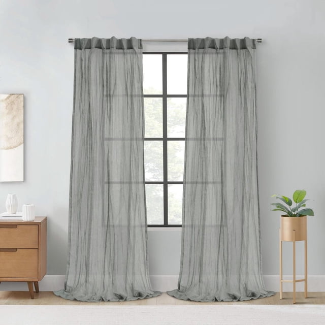 Loft Living Peoria Sheer Dual Header Single Curtain Panel 52 x 63 in Grey hanging on a metal rod over a window with black grid panes, made of 100% polyester sheer fabric with vertical texture and relaxed hems pooling on a light wood floor, in a neutral room with a wooden side table and white decor on the left and a gold planter with green plant on the right, designed to filter natural light while maintaining daytime privacy and suitable for home or office use.