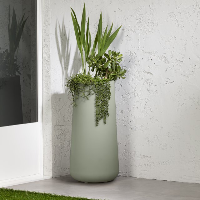 South Shore Dalya Tall Outdoor Pot Planter in sage green, a large cylindrical polyethylene resin planter with a matte finish, holding tall palm leaves and trailing succulents, placed on grey tile flooring against a textured white wall near a glass door, designed for outdoor use with weather, rust, and UV resistance, ideal for patios or balconies.