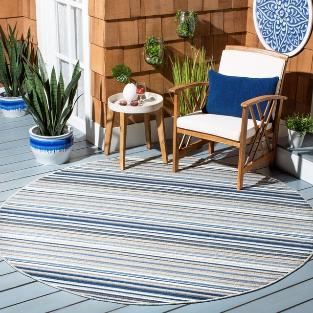 Safavieh Cabana Siomha Striped Outdoor Area Rug, round with blue, beige, and white horizontal stripes, on a grey wooden deck. Paired with a wooden armchair with white cushions and a blue pillow, a small round side table with decor, and surrounded by potted plants and a brown shingled wall with hanging planters. Ideal for outdoor patios or porches.